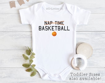 May include: White baby onesie with the text "NAP TIME" crossed out and "BASKETBALL" written below it. An orange basketball graphic is below the text. The onesie is on a white wooden background.