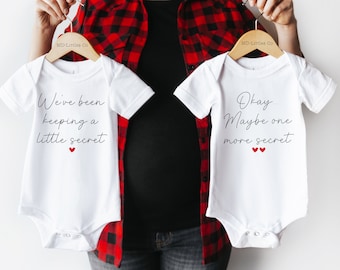 May include: Two white baby onesies with text. The onesie on the left says "We've been keeping a little secret". The onesie on the right says "Okay... Maybe one more secret".