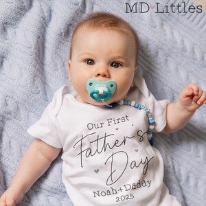 Op de afbeelding: Een witte romper met de tekst "Our First Father's Day Noah + Daddy 2025" erop. Een baby draagt de romper en ligt op een blauwe deken.