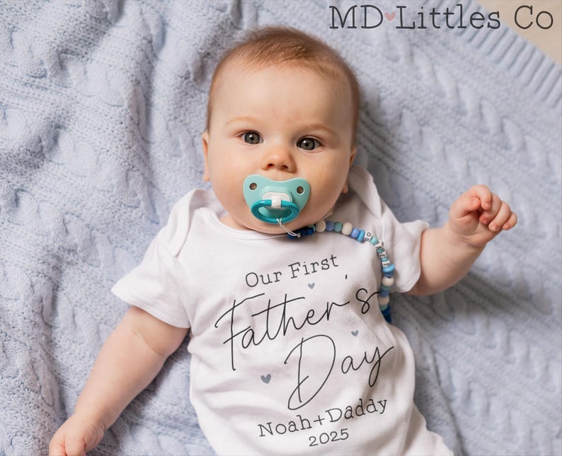 Op de afbeelding: Een witte romper met de tekst "Our First Father's Day Noah + Daddy 2025" erop. Een baby draagt de romper en ligt op een blauwe deken.