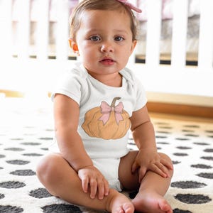 May include: A white baby onesie with a brown pumpkin with a pink bow on top. The onesie is on a baby sitting on a black and white polka dot rug.