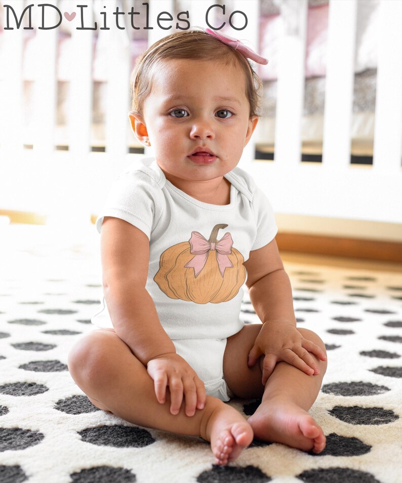 May include: A white baby onesie with a brown pumpkin with a pink bow on top. The onesie is on a baby sitting on a black and white polka dot rug.