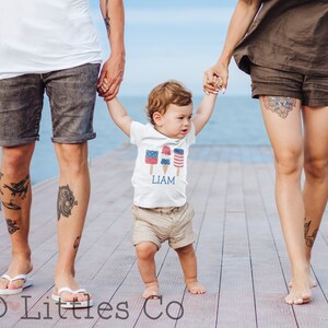 K&ouml;nnte beinhalten: Eine dreik&ouml;pfige Familie geht auf einem Holzsteg spazieren. Das Kind tr&auml;gt ein wei&szlig;es T-Shirt mit einem roten, wei&szlig;en und blauen Eiscreme-Motiv und dem Namen "LIAM" darauf gedruckt. Das Kind h&auml;lt beide Eltern an der Hand.