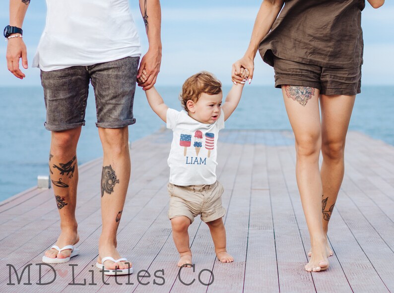 K&ouml;nnte beinhalten: Eine dreik&ouml;pfige Familie geht auf einem Holzsteg spazieren. Das Kind tr&auml;gt ein wei&szlig;es T-Shirt mit einem roten, wei&szlig;en und blauen Eiscreme-Motiv und dem Namen "LIAM" darauf gedruckt. Das Kind h&auml;lt beide Eltern an der Hand.