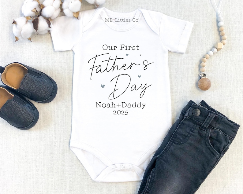 White onesie that says Our First Fathers Day in black with blue hearts around it. It has personalized names and a date at the bottom.