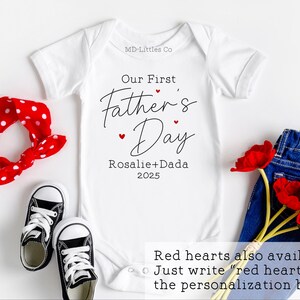 Op de afbeelding: Witte baby romper met de tekst "Our First Father's Day Rosalie+Dada 2025" in zwarte letters. Rode hartjes zijn rond de tekst verspreid. De tekst "Red hearts also available. Just write "red hearts" in the personalisation box." staat onderaan de afbeelding.