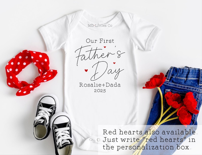 Op de afbeelding: Witte baby romper met de tekst "Our First Father's Day Rosalie+Dada 2025" in zwarte letters. Rode hartjes zijn rond de tekst verspreid. De tekst "Red hearts also available. Just write "red hearts" in the personalisation box." staat onderaan de afbeelding.