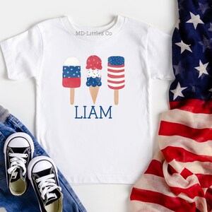 K&ouml;nnte beinhalten: Wei&szlig;es T-Shirt mit einem roten, wei&szlig;en und blauen Eiswaffel- und Eis am Stiel-Design. Der Text "LIAM" ist unter dem Eis gedruckt. Das Shirt liegt auf einer wei&szlig;en Oberfl&auml;che mit einer rot-wei&szlig;-blauen amerikanischen Flagge im Hintergrund.