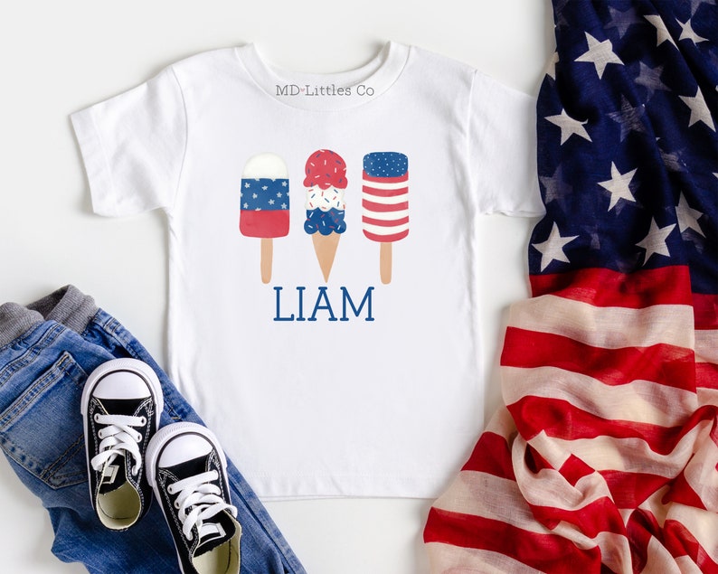 K&ouml;nnte beinhalten: Wei&szlig;es T-Shirt mit einem roten, wei&szlig;en und blauen Eiswaffel- und Eis am Stiel-Design. Der Text "LIAM" ist unter dem Eis gedruckt. Das Shirt liegt auf einer wei&szlig;en Oberfl&auml;che mit einer rot-wei&szlig;-blauen amerikanischen Flagge im Hintergrund.