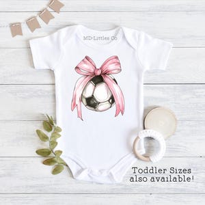 White onesie with a watercolor soccer ball. There is a large, pink bow on top of the ball.