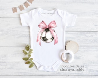 May include: White baby onesie with a pink bow and a soccer ball in the center. The text "MD-Littles Co" is printed on the onesie. The text "Toddler Sizes also available!" is printed below the onesie.