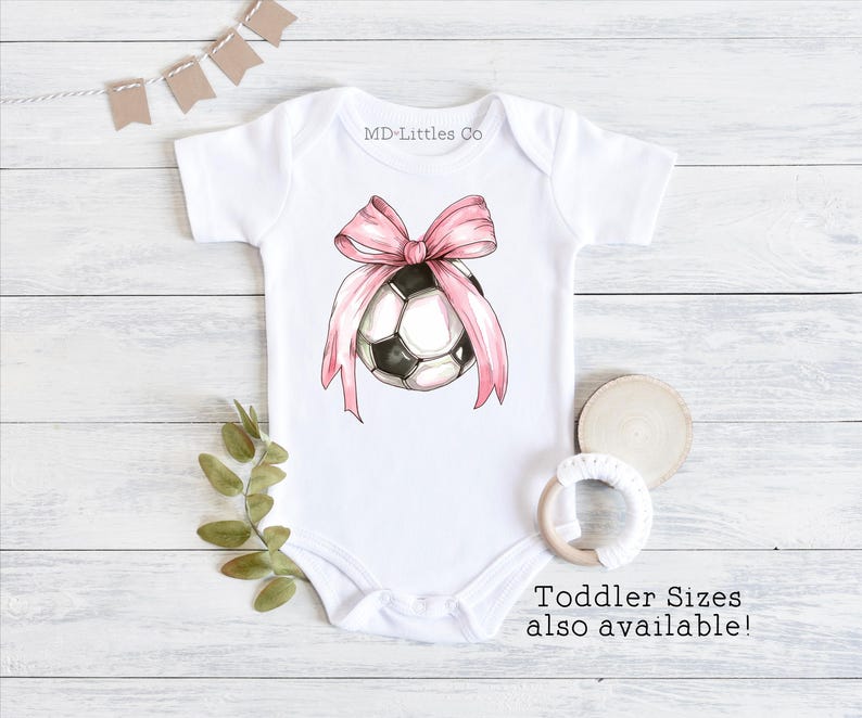 White onesie with a watercolor soccer ball. There is a large, pink bow on top of the ball.