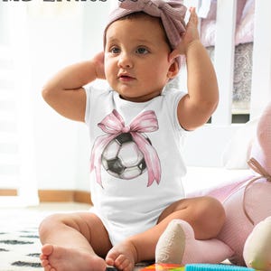 May include: A white baby onesie with a pink bow and a soccer ball design. The onesie is worn by a baby sitting on a pink and white blanket.