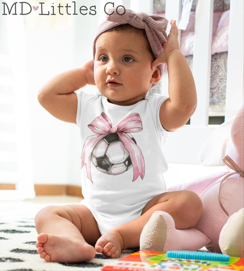 May include: A white baby onesie with a pink bow and a soccer ball design. The onesie is worn by a baby sitting on a pink and white blanket.