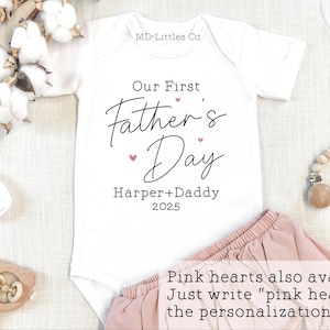 Op de afbeelding: Witte romper met de tekst "Our First Father's Day" in zwarte letters met roze hartjes. De tekst "Harper + Daddy 2025" is ook in zwart opgenomen. De romper ligt op een witte ondergrond met een roze rok en een houten speelgoed.