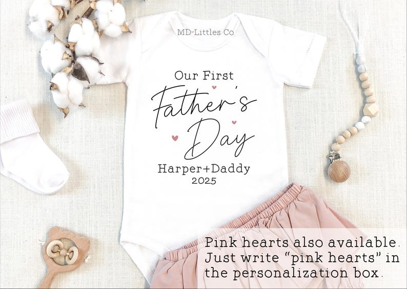 Op de afbeelding: Witte romper met de tekst "Our First Father's Day" in zwarte letters met roze hartjes. De tekst "Harper + Daddy 2025" is ook in zwart opgenomen. De romper ligt op een witte ondergrond met een roze rok en een houten speelgoed.