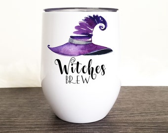 May include: A white stemless wine tumbler with a purple witch hat design and the words "Witches Brew" in black script. The tumbler is on a dark wood surface with a white fluffy background.