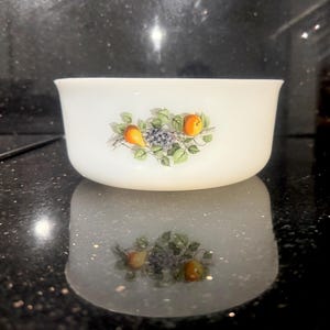 Könnte beinhalten: Weiße, ovale Schüssel mit einem Fruchtdesign. Die Schüssel zeigt eine Ansammlung von Birnen, Trauben und Orangen mit grünen Blättern. Die Schüssel steht auf einer reflektierenden Oberfläche.