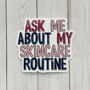 May include: A white sticker with the text "ASK ME ABOUT MY SKINCARE ROUTINE" in pink, dark blue, and burgundy lettering. The sticker is on a light wood grain surface.