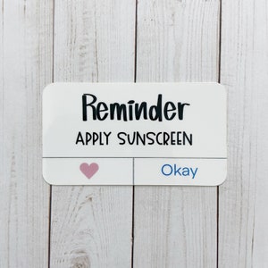 May include: White rectangular sticker with black text that reads "Reminder Apply Sunscreen". A pink heart is on the left side of the sticker and the word "Okay" is on the right side.