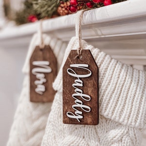 May include: Two white knitted stockings with wooden gift tags. The tags are brown and have the words "Mom" and "Daddy" written in white on them.