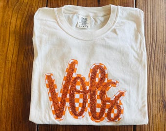 May include: A white t-shirt with an orange and white checkered "Vote" graphic.