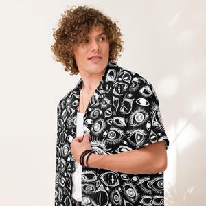 Puede incluir: Una camisa de botones de manga corta negra y blanca con un patrón repetitivo de ojos. La camisa tiene cuello y un corte holgado.
