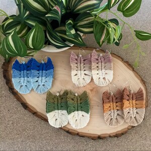 May include: Four pairs of handmade ombre macrame earrings. The earrings are in shades of blue, pink, green, and brown. The earrings are displayed on a wooden surface.