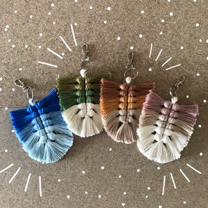 May include: Five colorful macrame keychains with fringe. The keychains are made with a variety of colors, including blue, green, brown, and white. Each keychain has a silver metal ring.