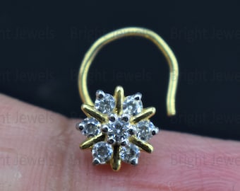 May include: A gold-colored nose ring with a flower-shaped design. The flower has a silver-colored center and white gemstones. The nose ring is held between two fingers against a dark background.