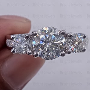 Puede incluir: Un anillo de compromiso de tres piedras con un gran diamante central y dos diamantes más pequeños a cada lado. El anillo está engastado en oro blanco y tiene un acabado pulido.
