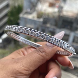 May include: A silver bracelet with a row of round, clear gemstones. The bracelet is held in a hand with a blurred background of a building and greenery.