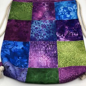 May include: A patchwork drawstring bag featuring a variety of colourful floral and abstract patterns in shades of green, blue, and purple. The bag has a cream-coloured rope drawstring closure and a gold button detail.