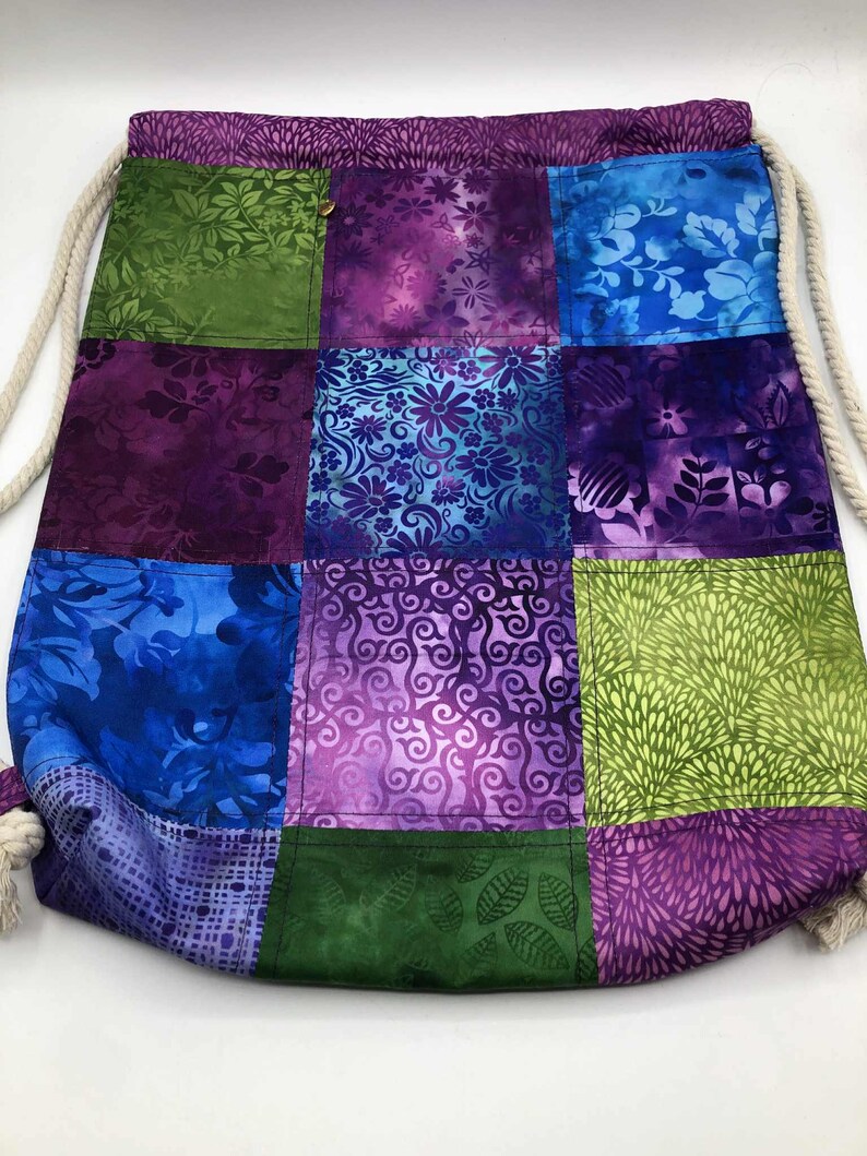 May include: A patchwork drawstring bag featuring a variety of colourful floral and abstract patterns in shades of green, blue, and purple. The bag has a cream-coloured rope drawstring closure and a gold button detail.