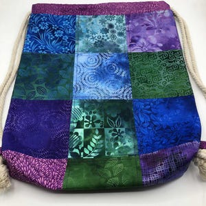 May include: A patchwork drawstring bag with a variety of blue, green, and purple floral and leaf patterns. The bag has a cream rope drawstring closure and a purple top and bottom trim.
