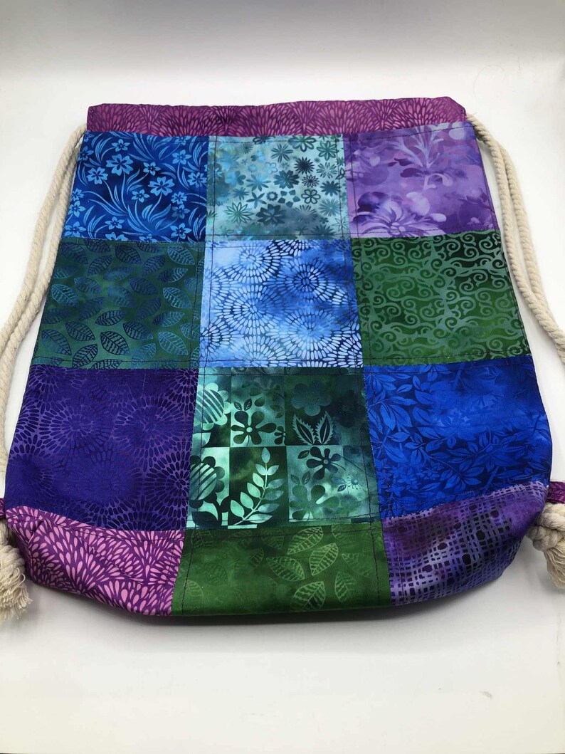 May include: A patchwork drawstring bag with a variety of blue, green, and purple floral and leaf patterns. The bag has a cream rope drawstring closure and a purple top and bottom trim.