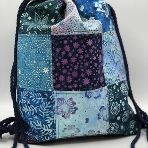 May include: A patchwork drawstring backpack in shades of blue, teal, and purple. The bag features a variety of floral and star patterns, with a dark blue rope drawstring. The bag is made of fabric.