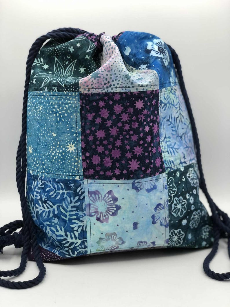 May include: A patchwork drawstring backpack in shades of blue, teal, and purple. The bag features a variety of floral and star patterns, with a dark blue rope drawstring. The bag is made of fabric.