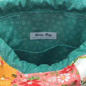 K&ouml;nnte beinhalten: Eine gr&uuml;ne Stofftasche mit wei&szlig;en Punkten. Die Tasche hat einen Kordelzugverschluss und ein Etikett mit der Aufschrift "Nonna's Bags Made in New Hampshire". Die Tasche ist mit einem Blumenstoff gef&uuml;ttert.