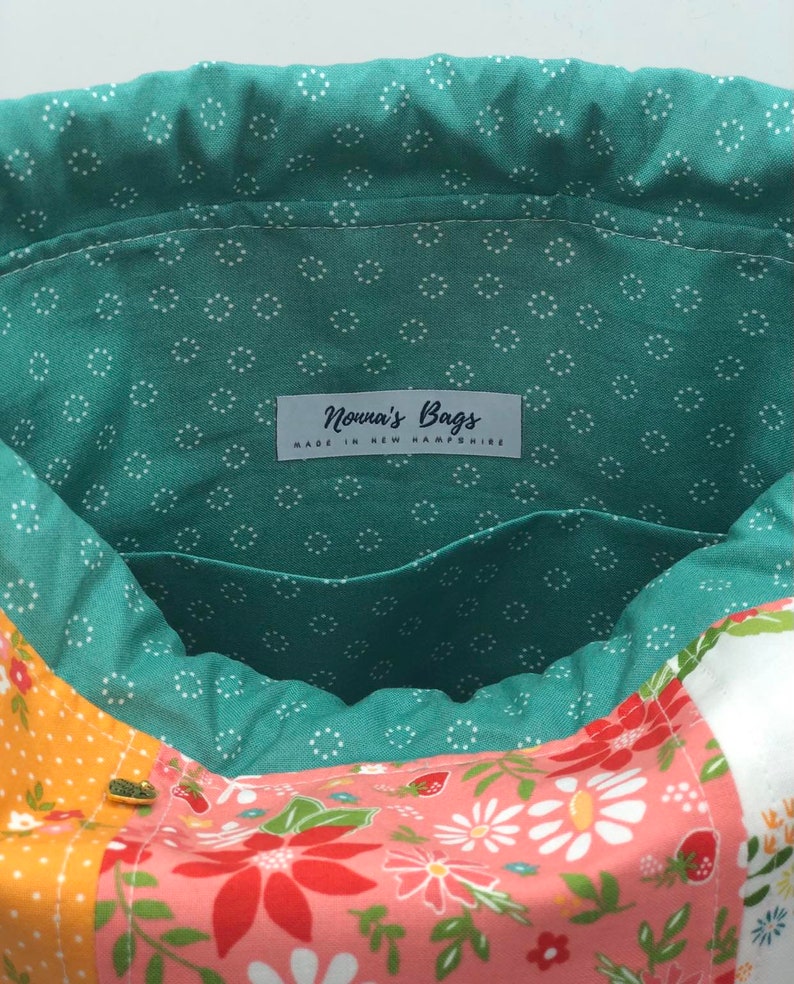 K&ouml;nnte beinhalten: Eine gr&uuml;ne Stofftasche mit wei&szlig;en Punkten. Die Tasche hat einen Kordelzugverschluss und ein Etikett mit der Aufschrift "Nonna's Bags Made in New Hampshire". Die Tasche ist mit einem Blumenstoff gef&uuml;ttert.