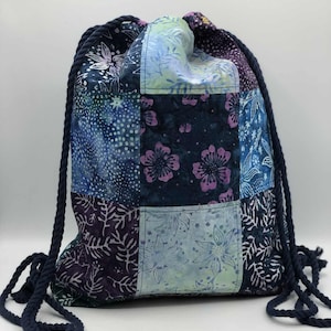 May include: A patchwork drawstring backpack with a navy blue rope drawstring. The bag features a variety of blue, purple, and green floral patterned fabric squares. The bag is designed for carrying items.