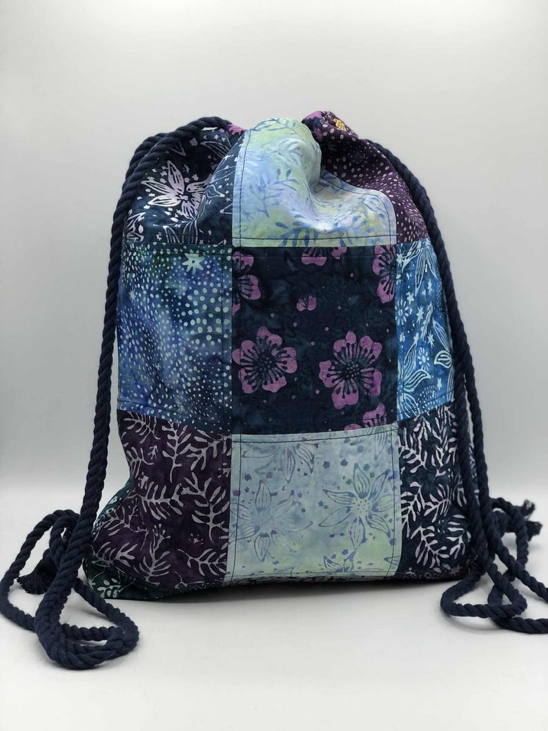 May include: A patchwork drawstring backpack with a navy blue rope drawstring. The bag features a variety of blue, purple, and green floral patterned fabric squares. The bag is designed for carrying items.