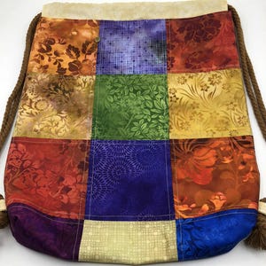 以下が含まれることがあります： 様々な色とりどりの花柄や模様の生地を使ったパッチワーク巾着袋。ベージュのトップと、タッセル付きの茶色のロープのドローストリングが特徴です。オレンジ、赤、緑、青、黄色の色合いの正方形の生地で作られています。