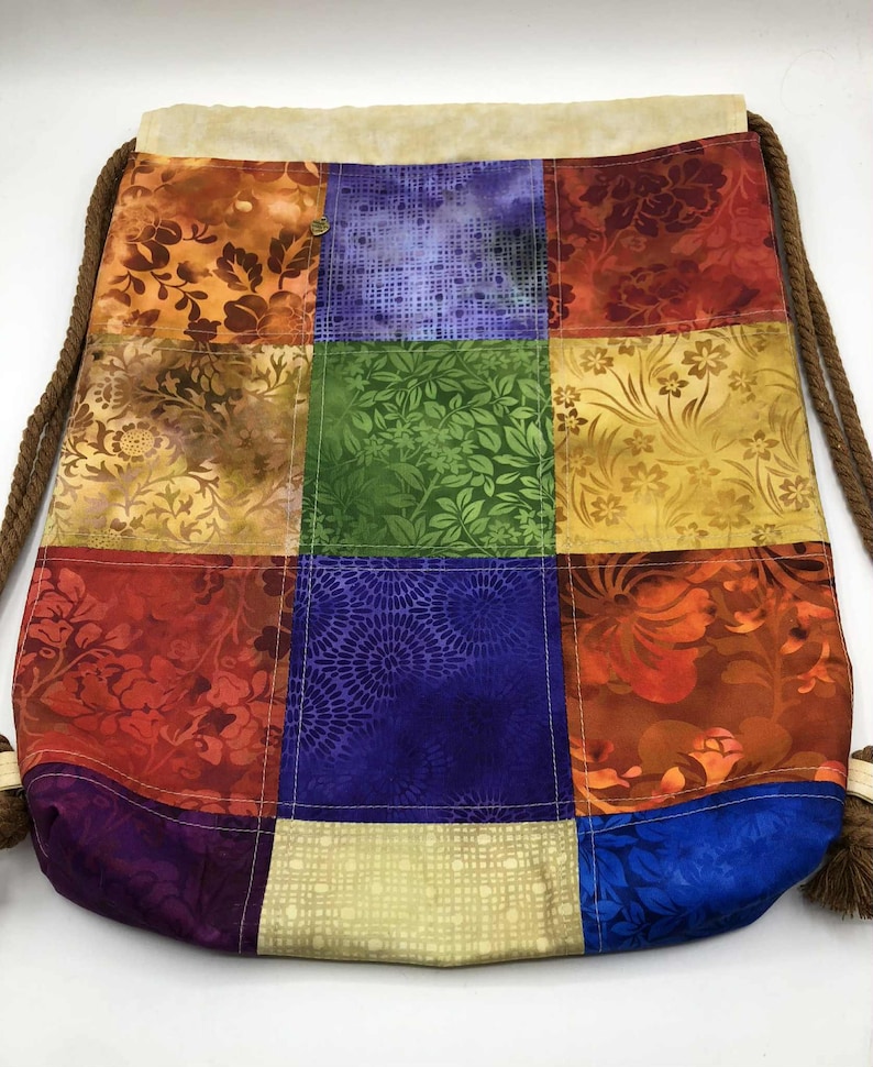 以下が含まれることがあります： 様々な色とりどりの花柄や模様の生地を使ったパッチワーク巾着袋。ベージュのトップと、タッセル付きの茶色のロープのドローストリングが特徴です。オレンジ、赤、緑、青、黄色の色合いの正方形の生地で作られています。