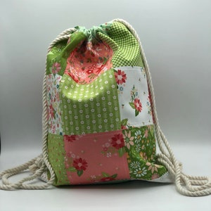 May include: A green and pink patchwork drawstring bag with white rope handles. The bag is made of different floral patterned fabrics.