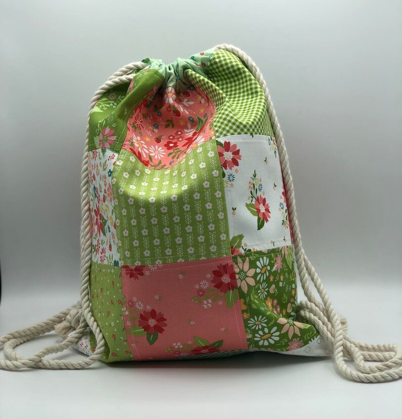 May include: A green and pink patchwork drawstring bag with white rope handles. The bag is made of different floral patterned fabrics.