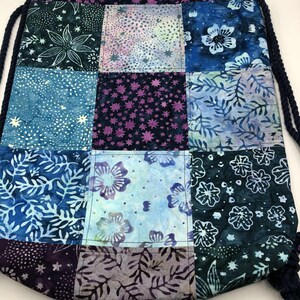 May include: A patchwork drawstring bag with a variety of blue, purple, and white floral and star patterns. The bag has a dark blue drawstring and a matching patterned top border. The bag is made of fabric.