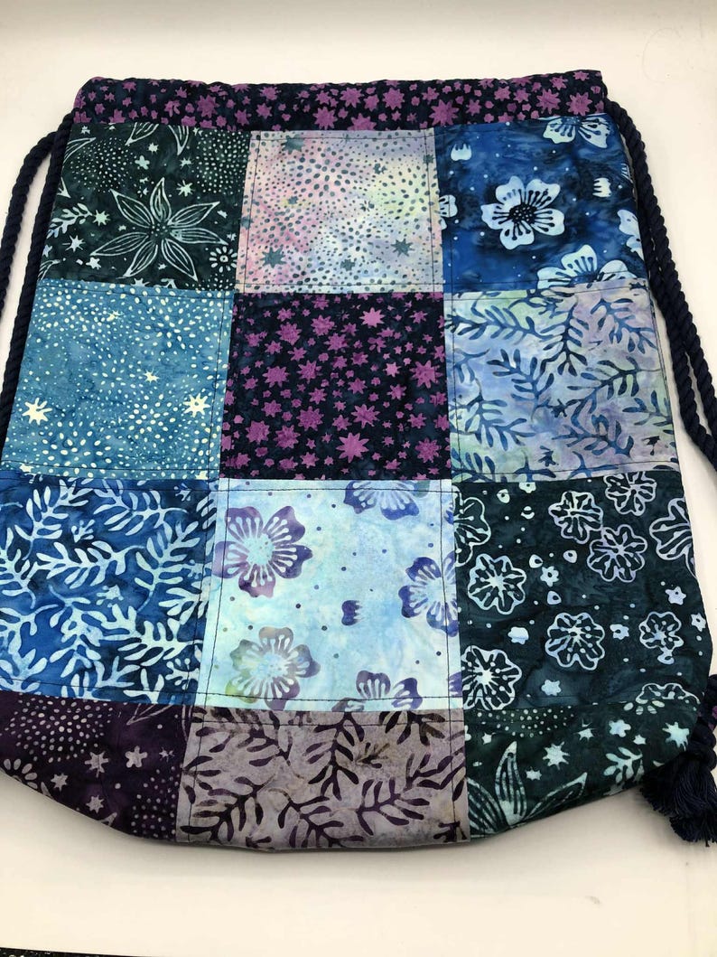 May include: A patchwork drawstring bag with a variety of blue, purple, and white floral and star patterns. The bag has a dark blue drawstring and a matching patterned top border. The bag is made of fabric.