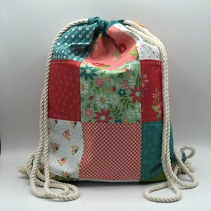 K&ouml;nnte beinhalten: Eine Patchwork-Kordeltasche mit einem wei&szlig;en Seilgriff. Die Tasche besteht aus verschiedenen Stoffen mit Blumen-, Punkte- und Karomustern in Rot-, Gr&uuml;n- und Wei&szlig;t&ouml;nen.