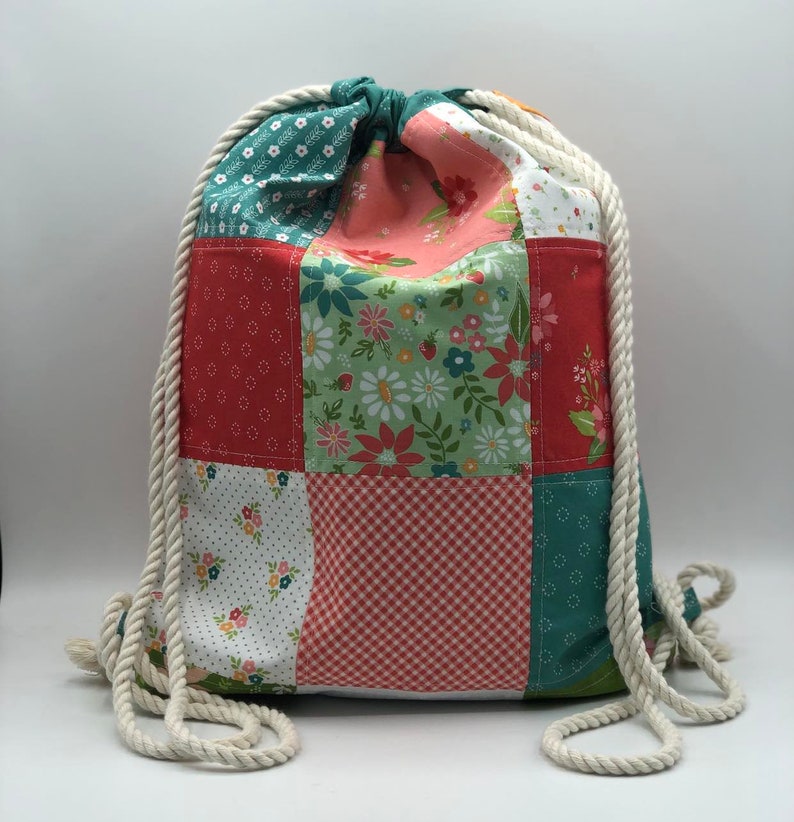 K&ouml;nnte beinhalten: Eine Patchwork-Kordeltasche mit einem wei&szlig;en Seilgriff. Die Tasche besteht aus verschiedenen Stoffen mit Blumen-, Punkte- und Karomustern in Rot-, Gr&uuml;n- und Wei&szlig;t&ouml;nen.